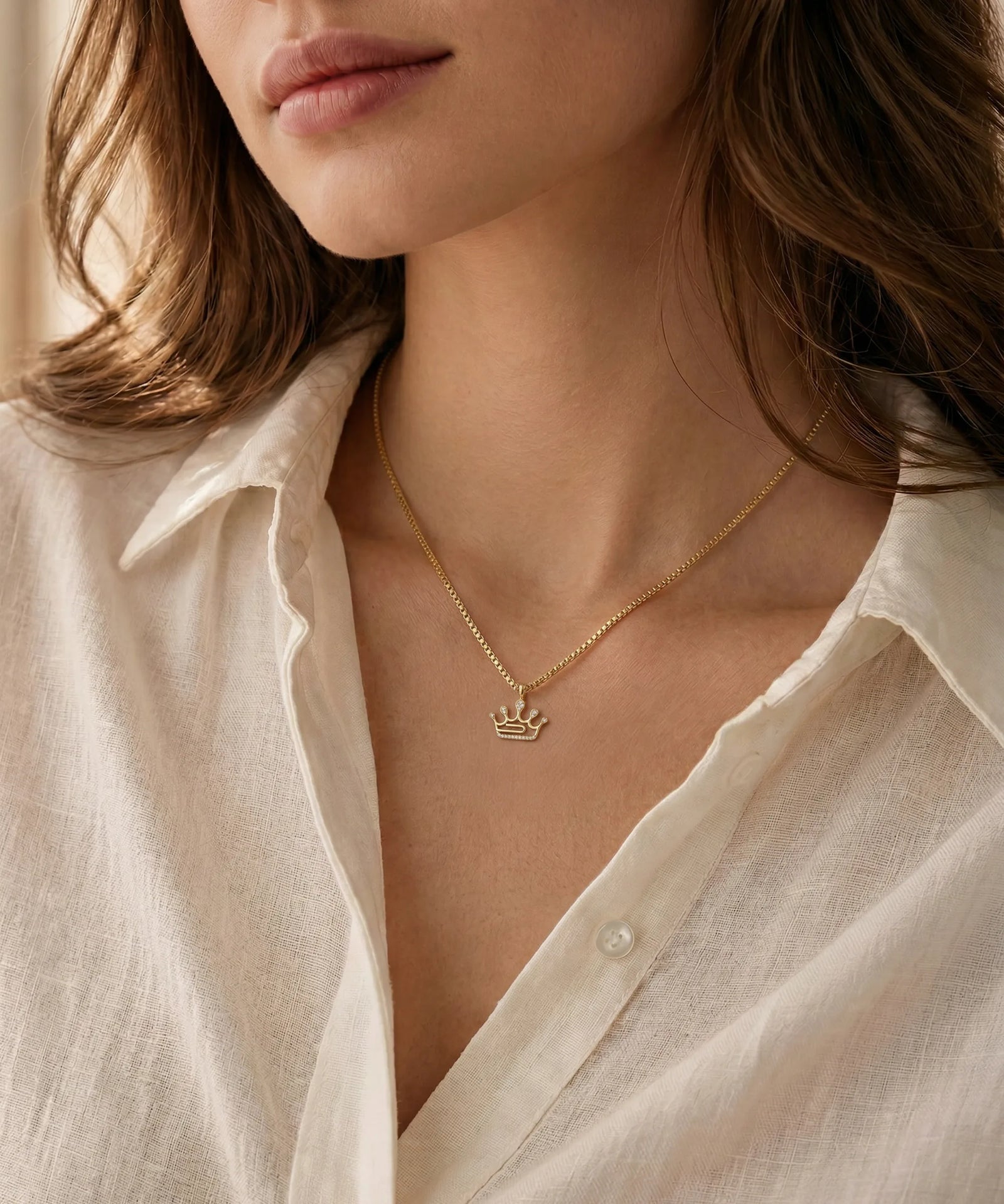 Close-up of a person wearing a gold necklace with a crown pendant, against a neutral background.