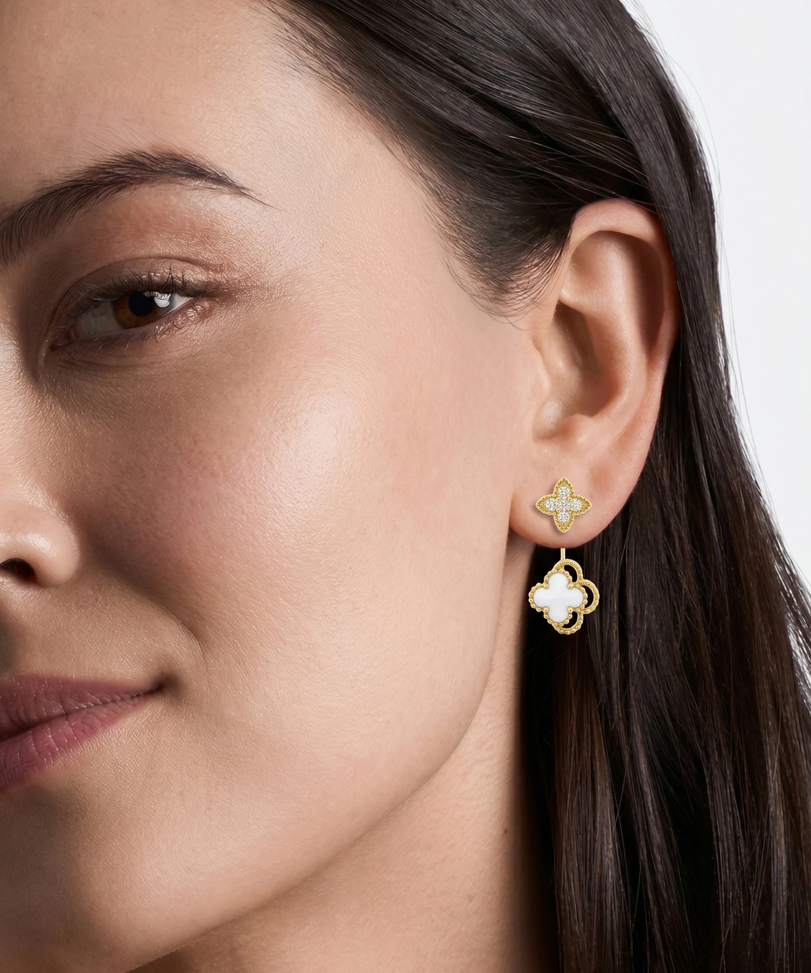 A close-up of a woman's ear wearing a pair of gold and white clover-shaped drop earrings against a clean light gray background.