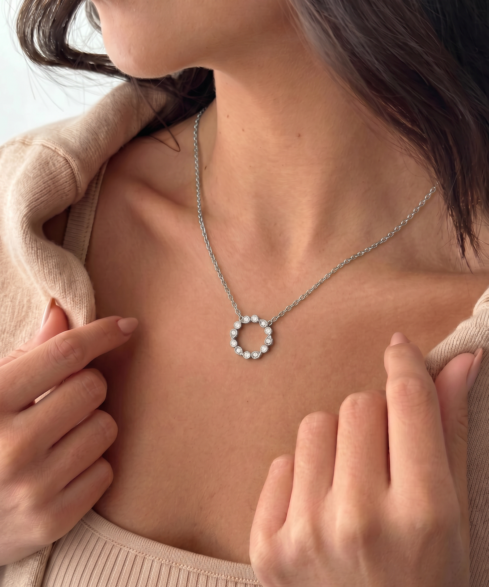 A close-up lifestyle shot of a woman wearing a delicate silver chain necklace with a small circular pendant adorned with tiny clear stones. She is wearing a beige knit top, and her hands are gently touching the fabric near her shoulders.