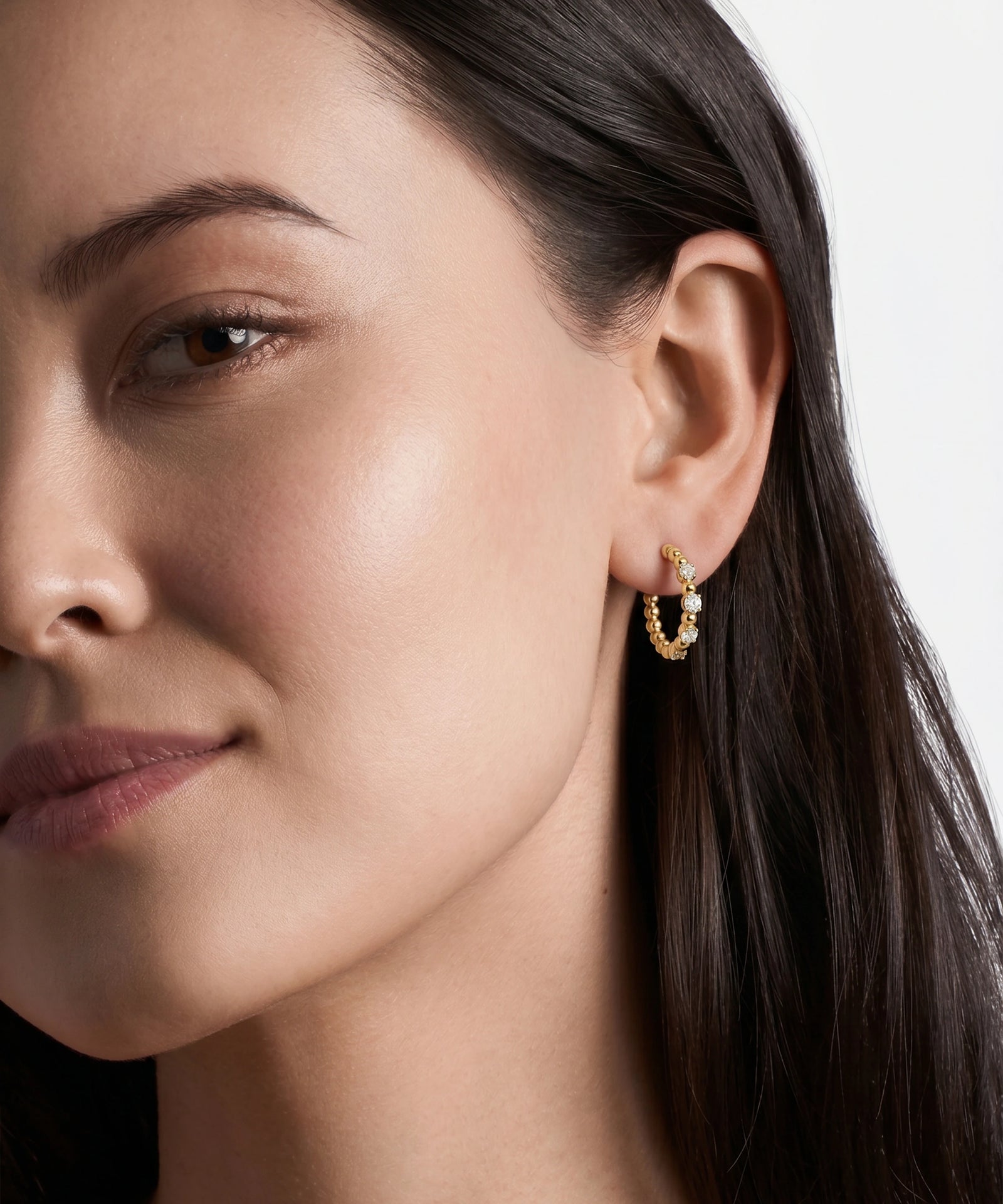 A close-up of a woman's ear wearing a small gold crescent hoop earring with tiny clear stones, against a clean light gray background.