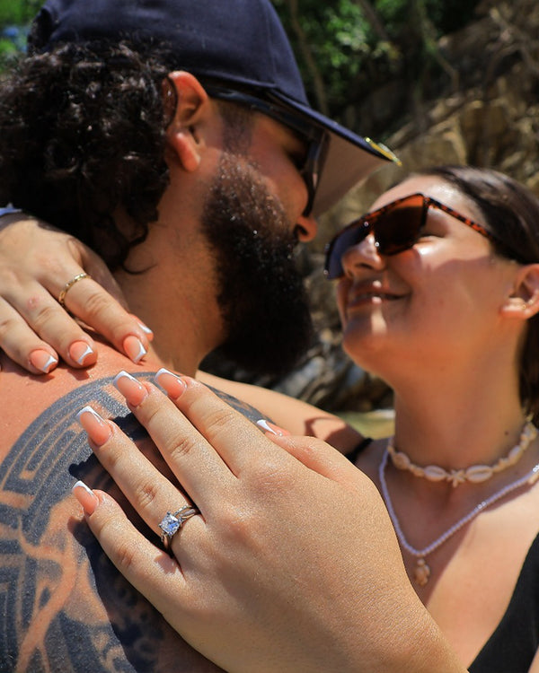 A couple embraces outdoors, smiling at each other. The woman wears sunglasses and a shell necklace, and her hand, featuring a diamond engagement ring, rests on the man’s tattooed shoulder.
