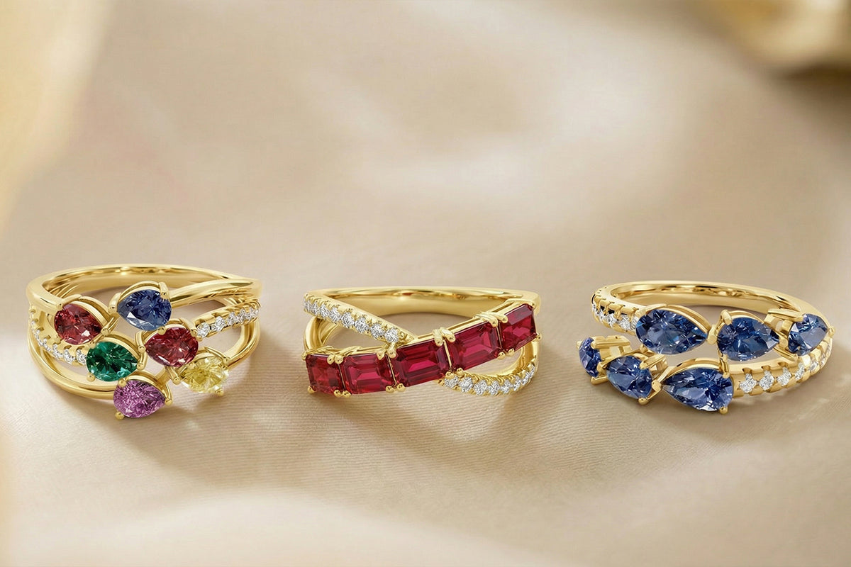 Three gold rings on a beige background. The left ring has multiple colored gemstones. The middle ring has red square gemstones and diamonds. The right ring has blue pear-shaped gemstones and diamonds.