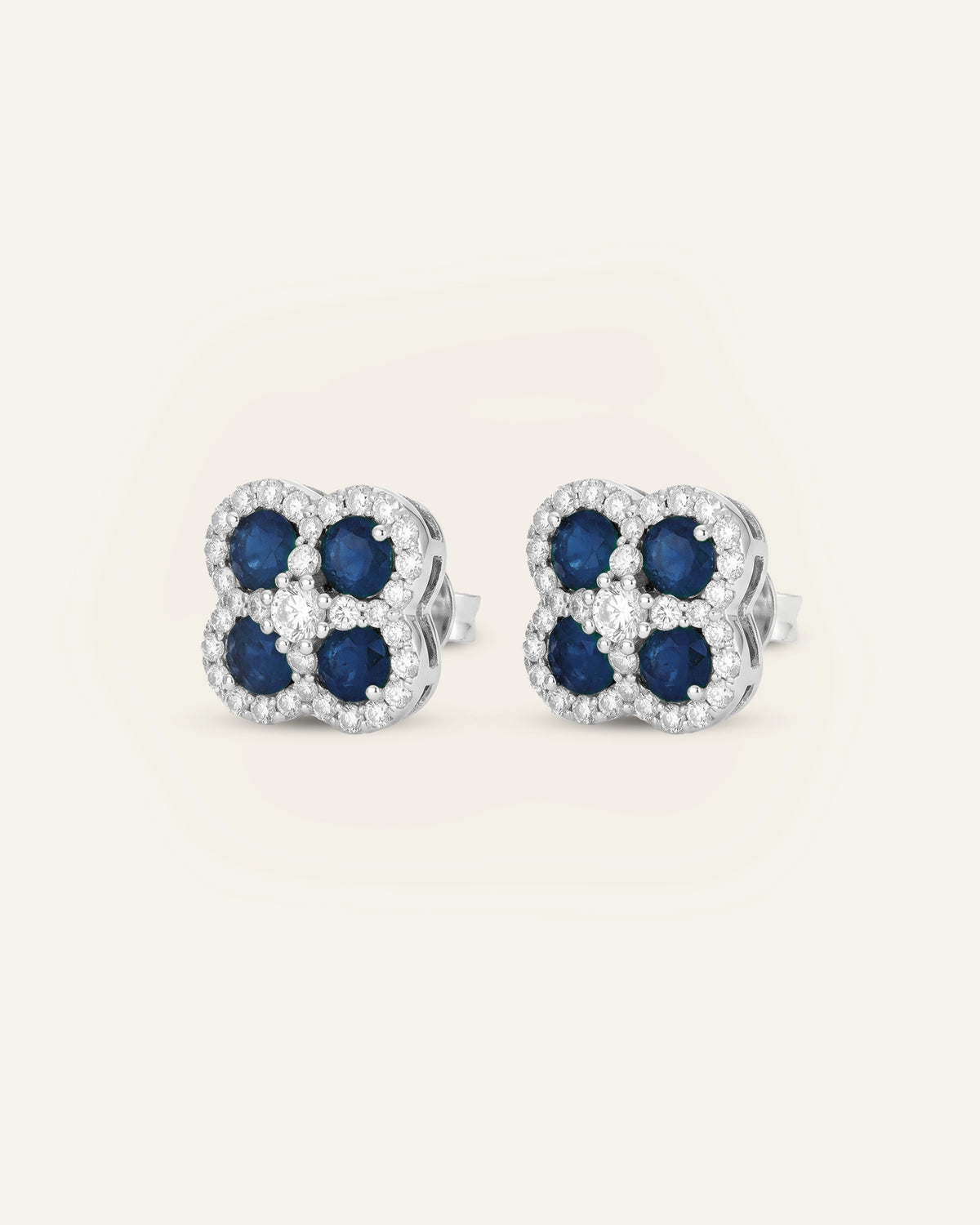 A pair of silver-toned stud earrings featuring a four-petal clover design with dark blue round-cut center stones surrounded by clear pavé-set stones, shown on a light cream background.
