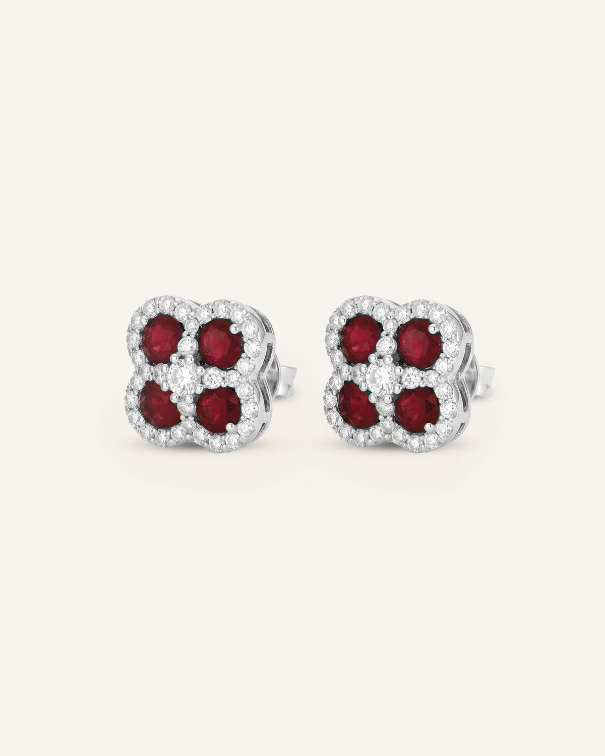 A pair of silver-toned stud earrings featuring four round-cut deep red gemstones in a four-petal cluster setting, surrounded by a border of small clear stones, shown on a light cream background.