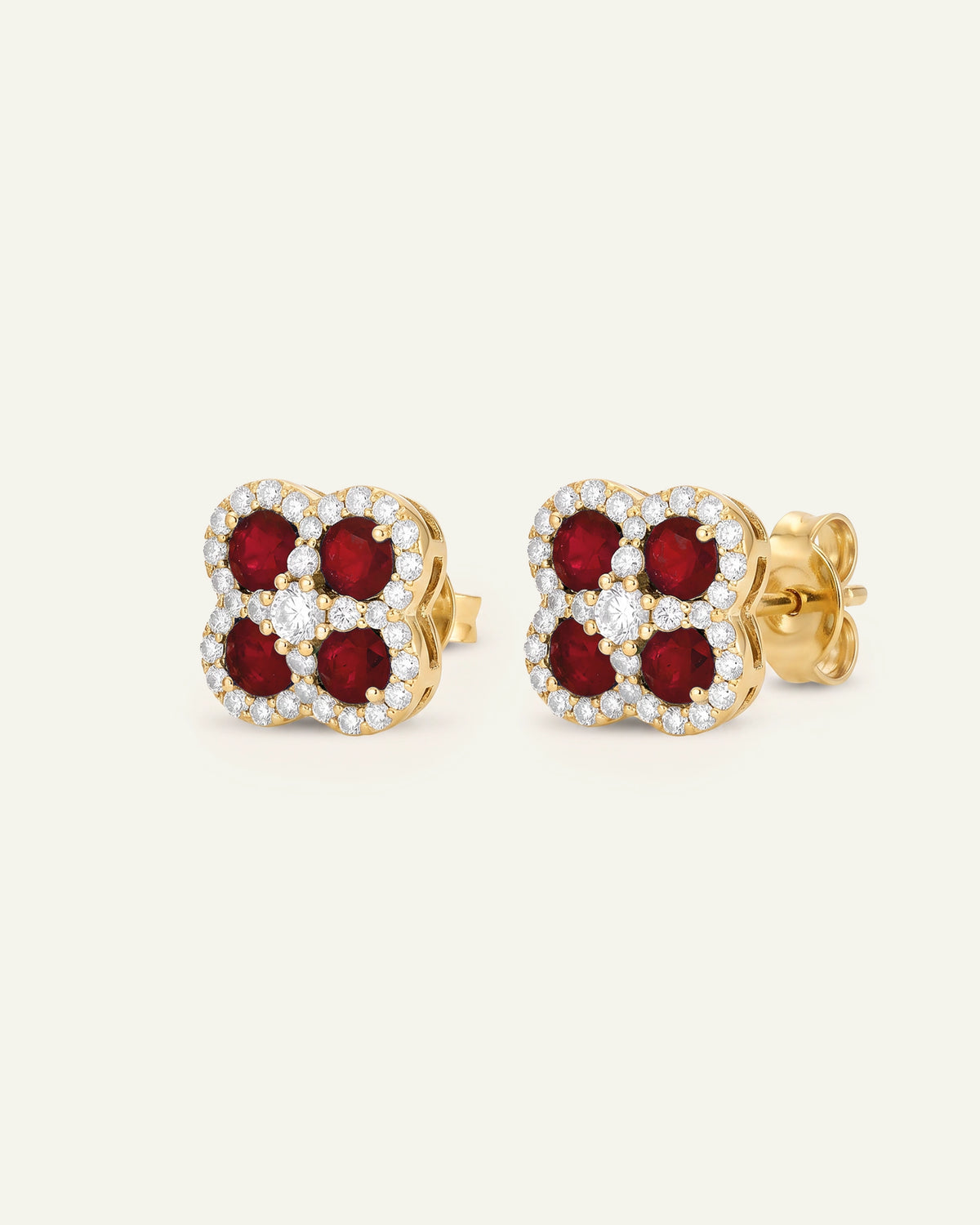 A pair of gold four-petal stud earrings featuring deep red gemstones and a halo of small clear stones, shown on a light cream background.