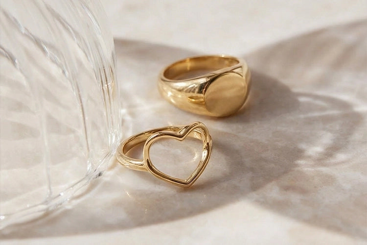 Two gold rings, one heart-shaped and one plain, on a light surface with a glass object.