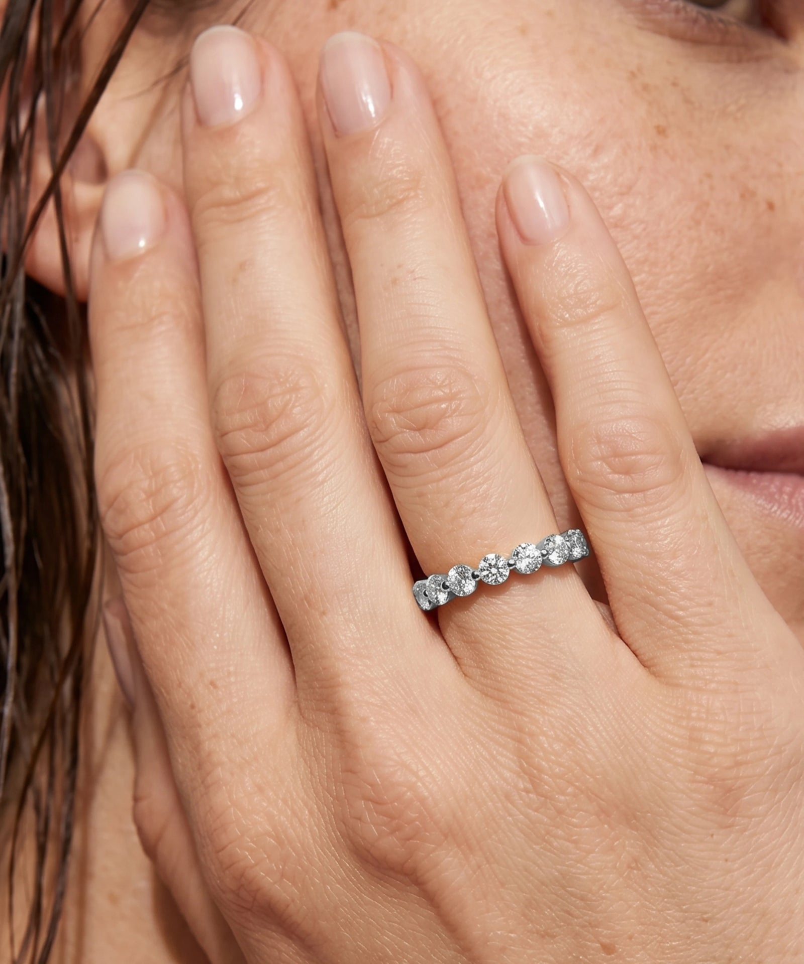 A close-up of a person's hand wearing a silver-toned diamond eternity ring, with the hand resting against their face.