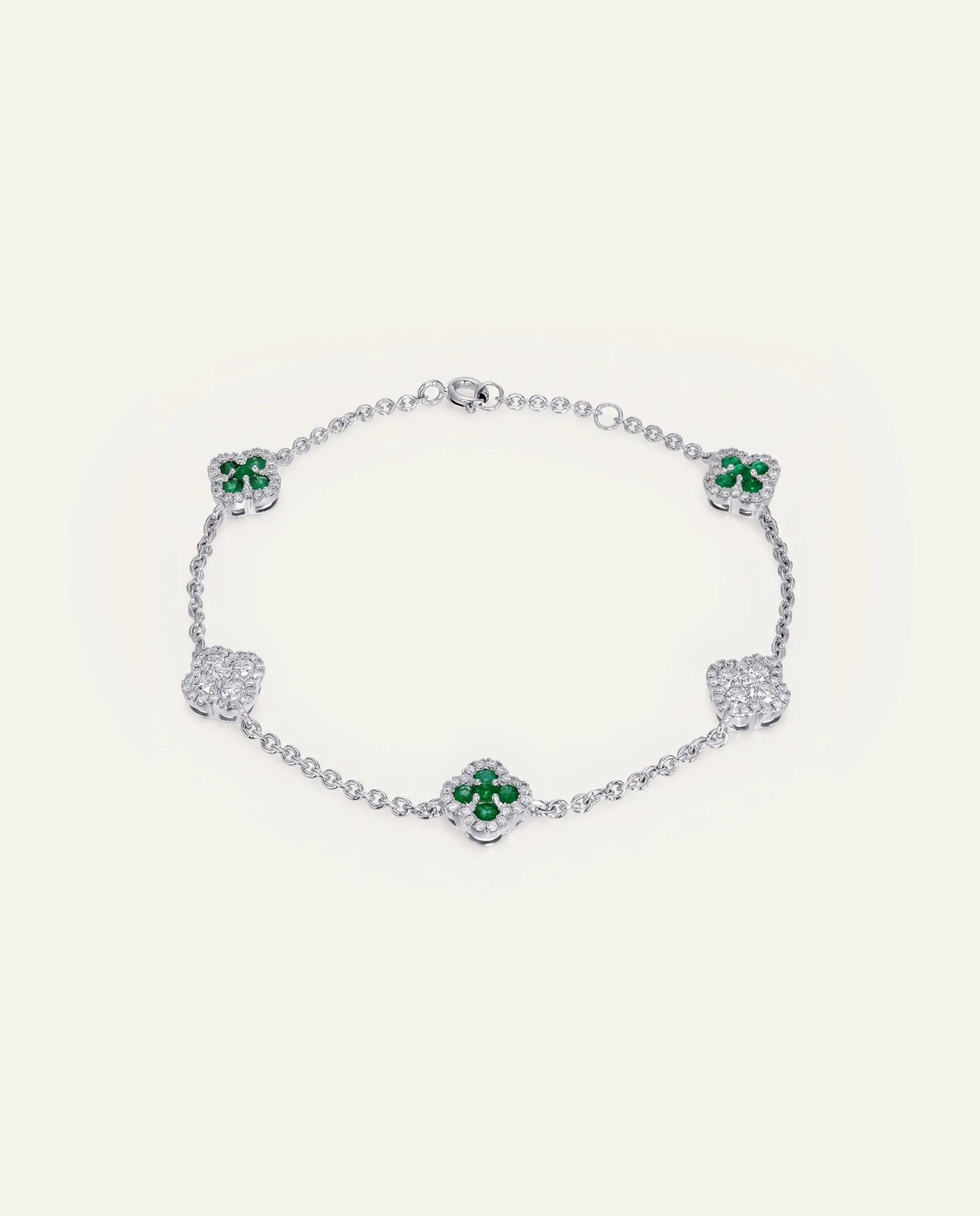 A silver-toned chain bracelet featuring five clover motifs, with three clovers accented by vibrant green gemstones and two larger clovers paved with clear stones, set against a solid off-white background.