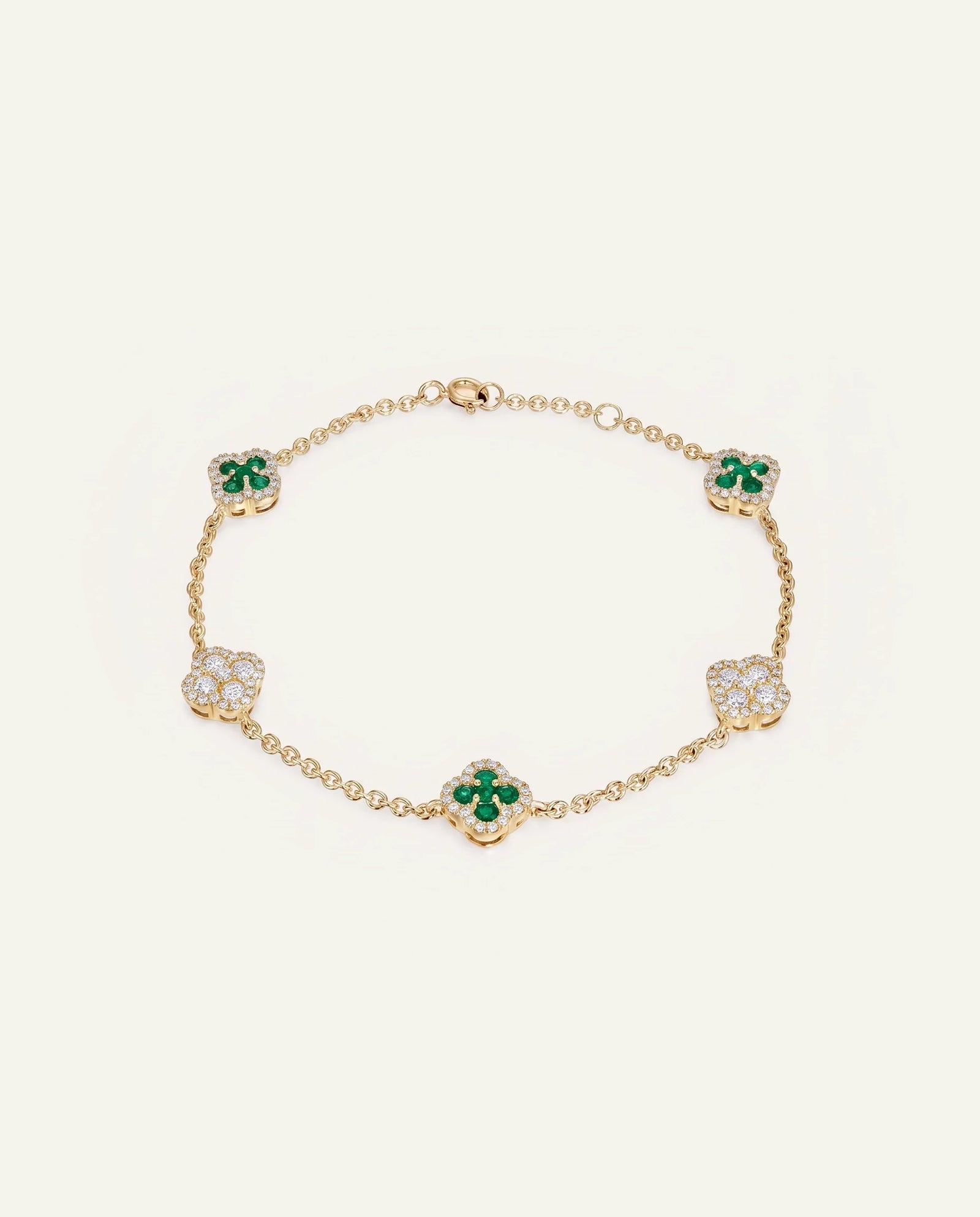 A delicate gold chain bracelet featuring five clover motifs, with three clovers set with vibrant green emeralds and two set with clear pavé diamonds, displayed on a clean off-white background.