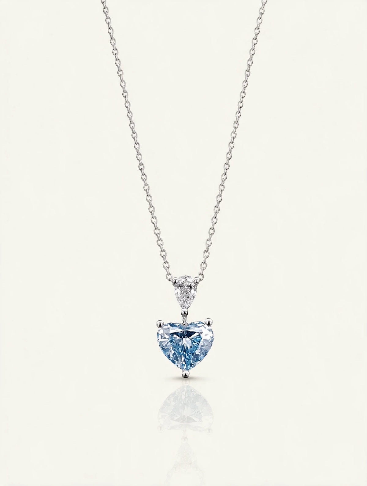 A delicate silver chain necklace featuring a heart-shaped light blue gemstone pendant in a silver setting, displayed on a clean off-white background with a soft reflection below.