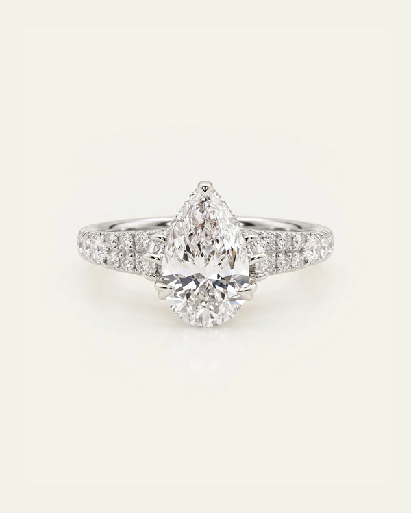 A pear-cut diamond engagement ring with a pavé band set with small round diamonds, shown on a light cream background.
