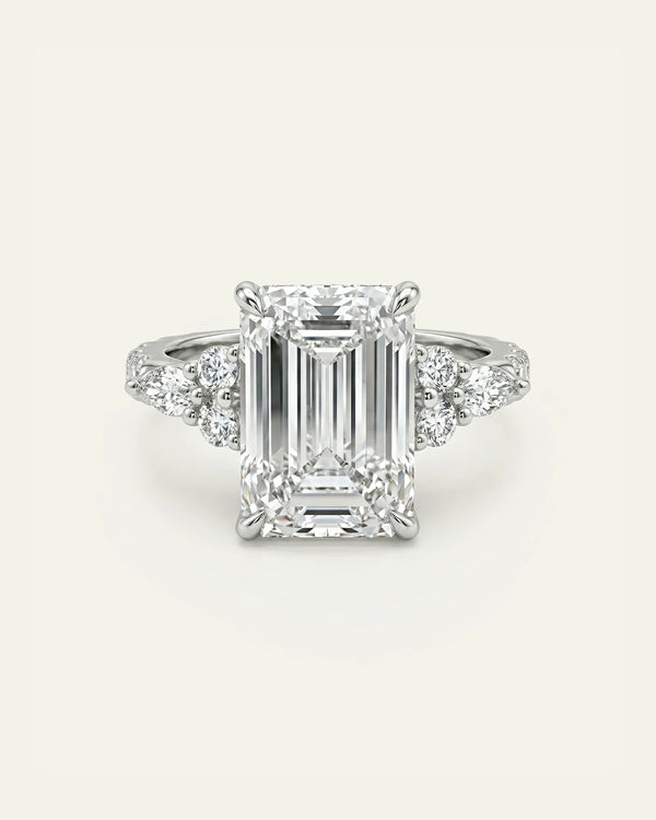 An emerald-cut diamond engagement ring with a silver-toned band featuring small side diamonds, centered on a light cream background.