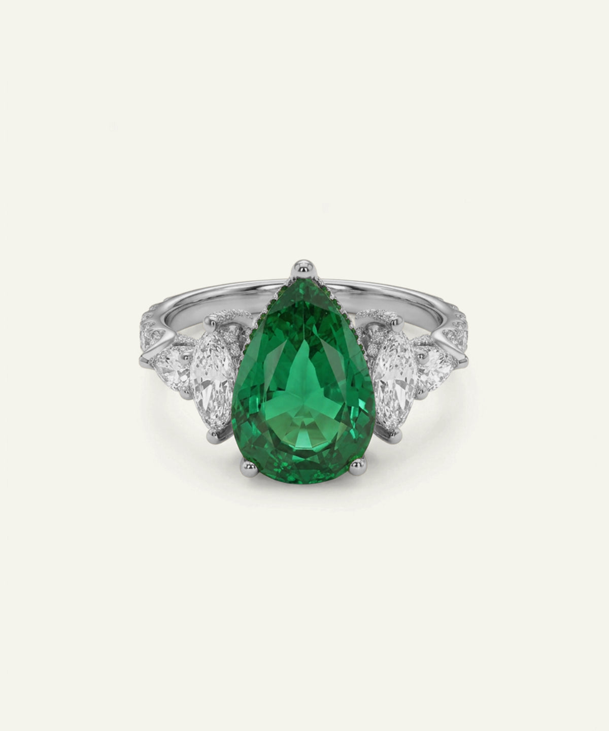 A pear-cut emerald green gemstone ring set in a silver-toned metal with diamond accents on the shoulders, centered on a light cream background.