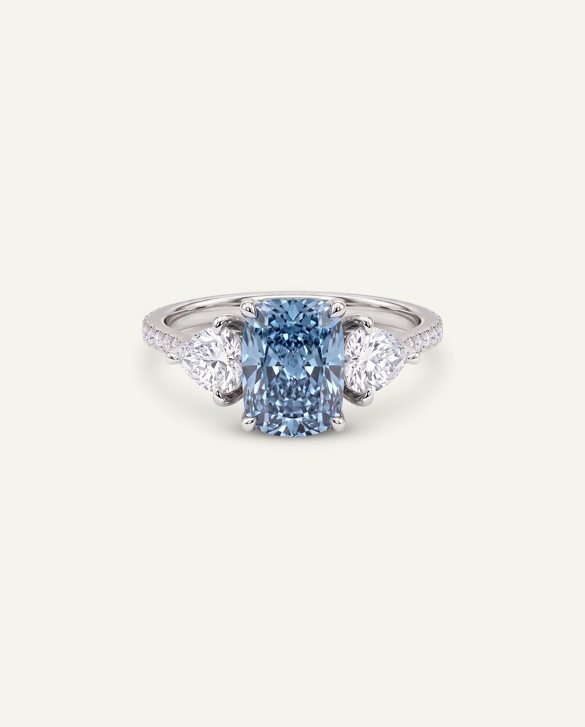 A three-stone diamond ring featuring a central cushion-cut blue diamond flanked by two round diamonds, set on a silver-colored band, shown against a solid off-white background.