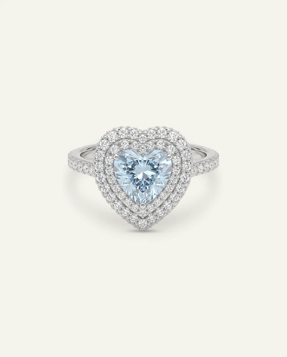 A silver ring featuring a heart-cut light blue diamond center stone surrounded by a double halo of round clear diamonds, with a pavé diamond band set against a light cream background.