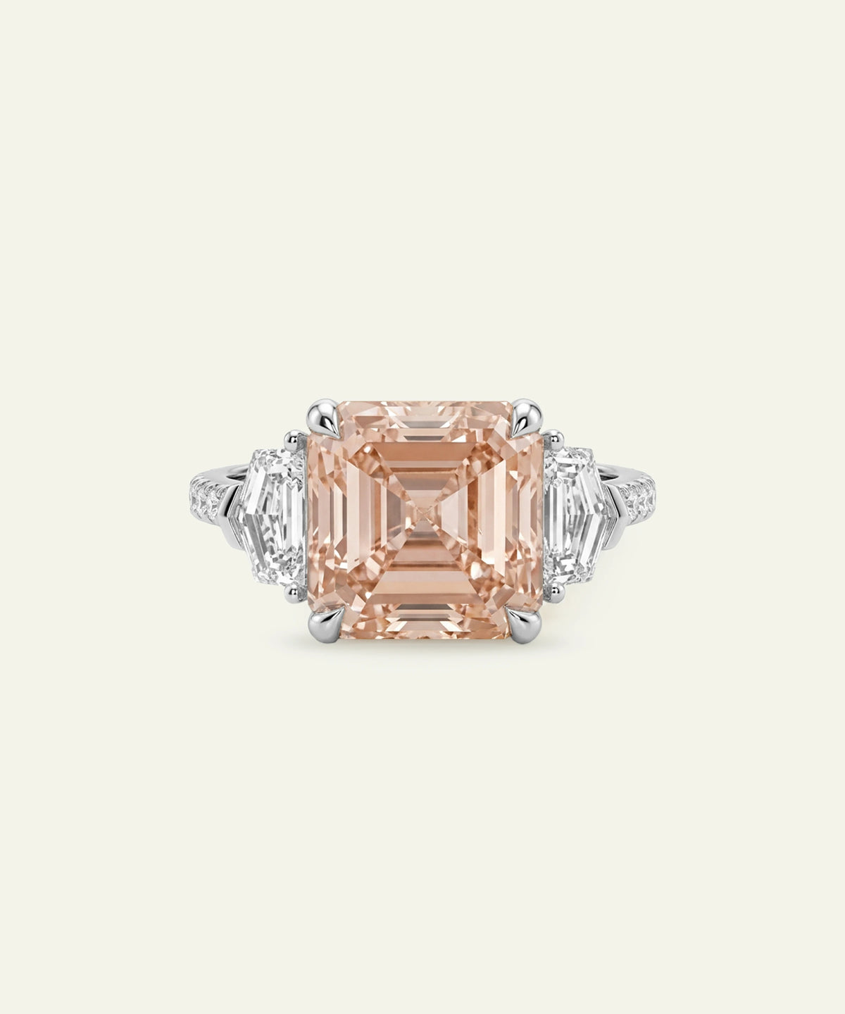 A silver ring featuring a large emerald-cut peach or champagne diamond center stone flanked by trapezoid-shaped side diamonds and a pavé-set diamond band, set against a light cream background.