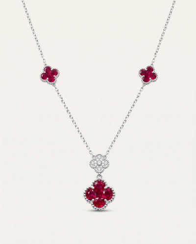 A silver-tone necklace with three floral clover motifs: two small clovers on the chain and one larger clover pendant set with deep red gemstones and small clear stones, shown on a light beige background. - alternate view