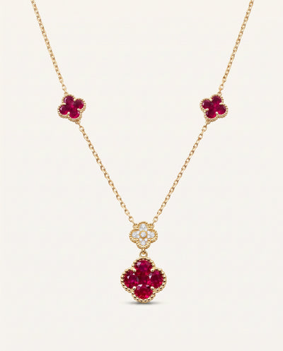 A gold necklace featuring three floral clover motifs: two small clovers on the chain and one larger clover pendant surrounded by clear stones, set against a light cream background.