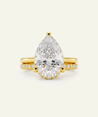 A pear-cut diamond ring with a yellow gold band featuring pavé-set diamonds, centered on a light cream background.