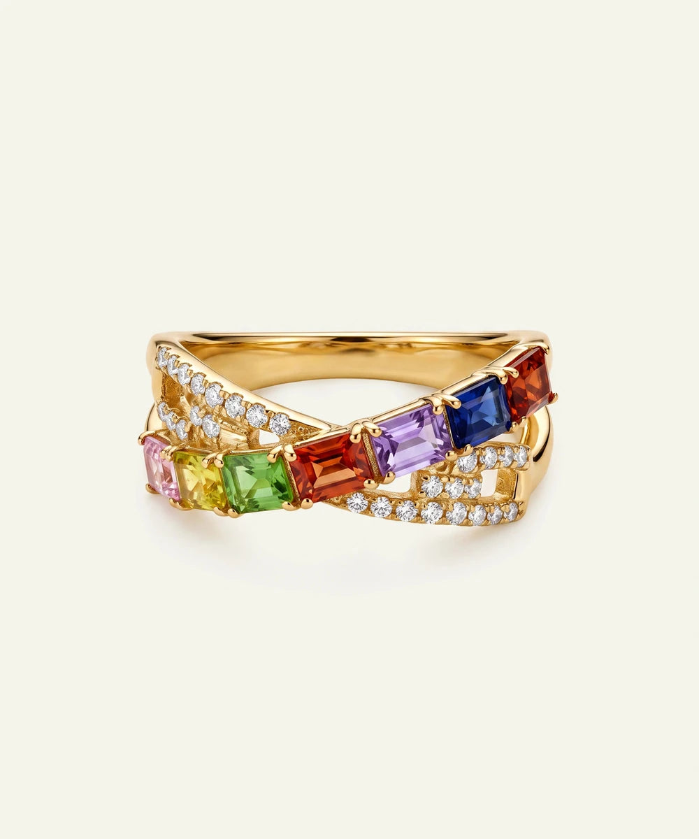 A gold ring featuring a double crossover design with baguette-cut rainbow gemstones and pavÃ©-set clear stones, shown on a light cream background.