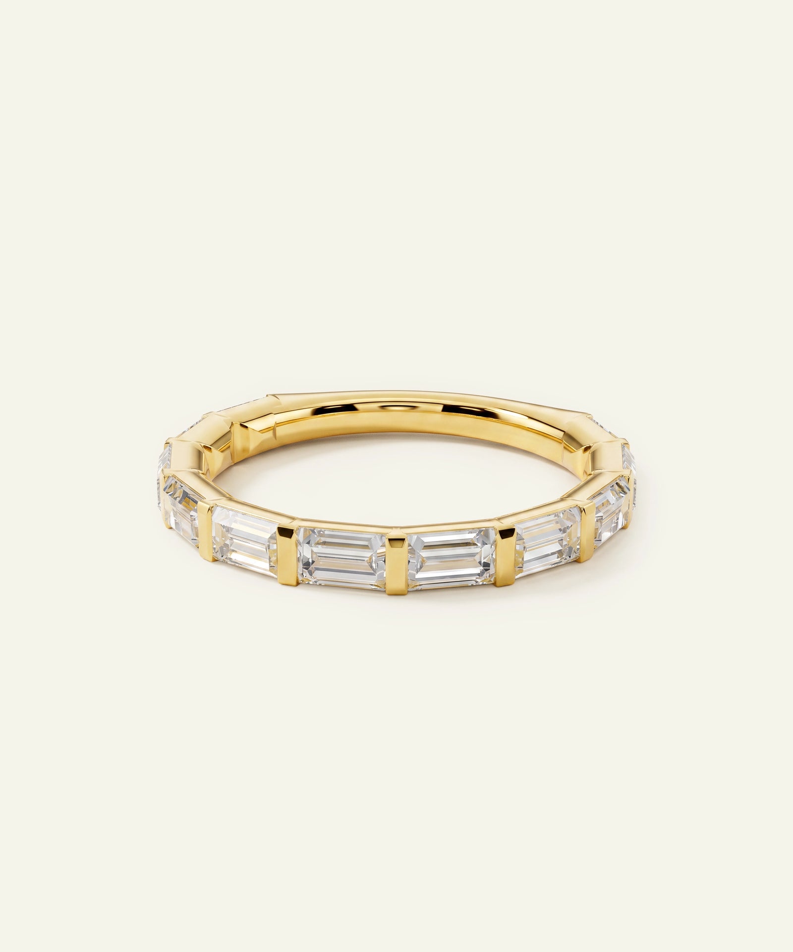 A gold band ring featuring a row of clear baguette-cut gemstones, photographed on a clean, solid off-white background.