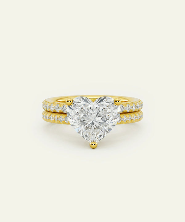 A yellow gold engagement ring featuring a large heart-shaped diamond center stone, flanked by two slim diamond-paved bands, set against a solid off-white background.