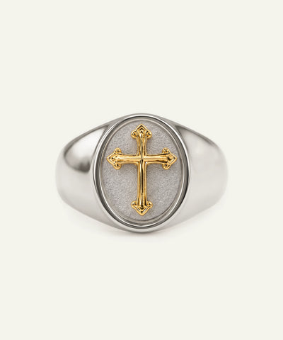 A silver signet ring featuring a polished gold cross inlaid on a textured, light gray oval face, set against a solid off-white background.