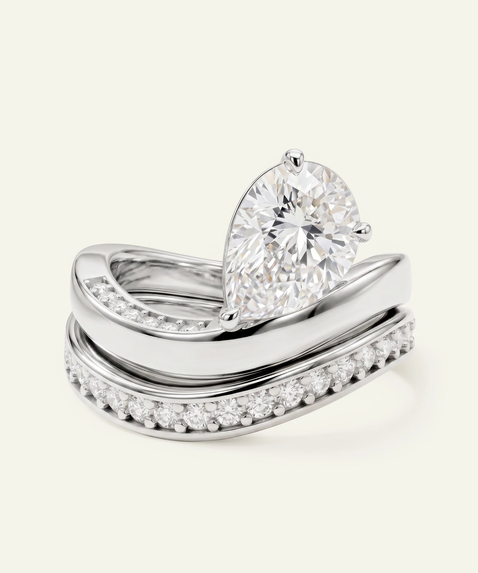 A silver-toned wedding ring set featuring a pear-cut diamond engagement ring stacked on top of a matching curved band with pavé diamonds, photographed on a light cream background.