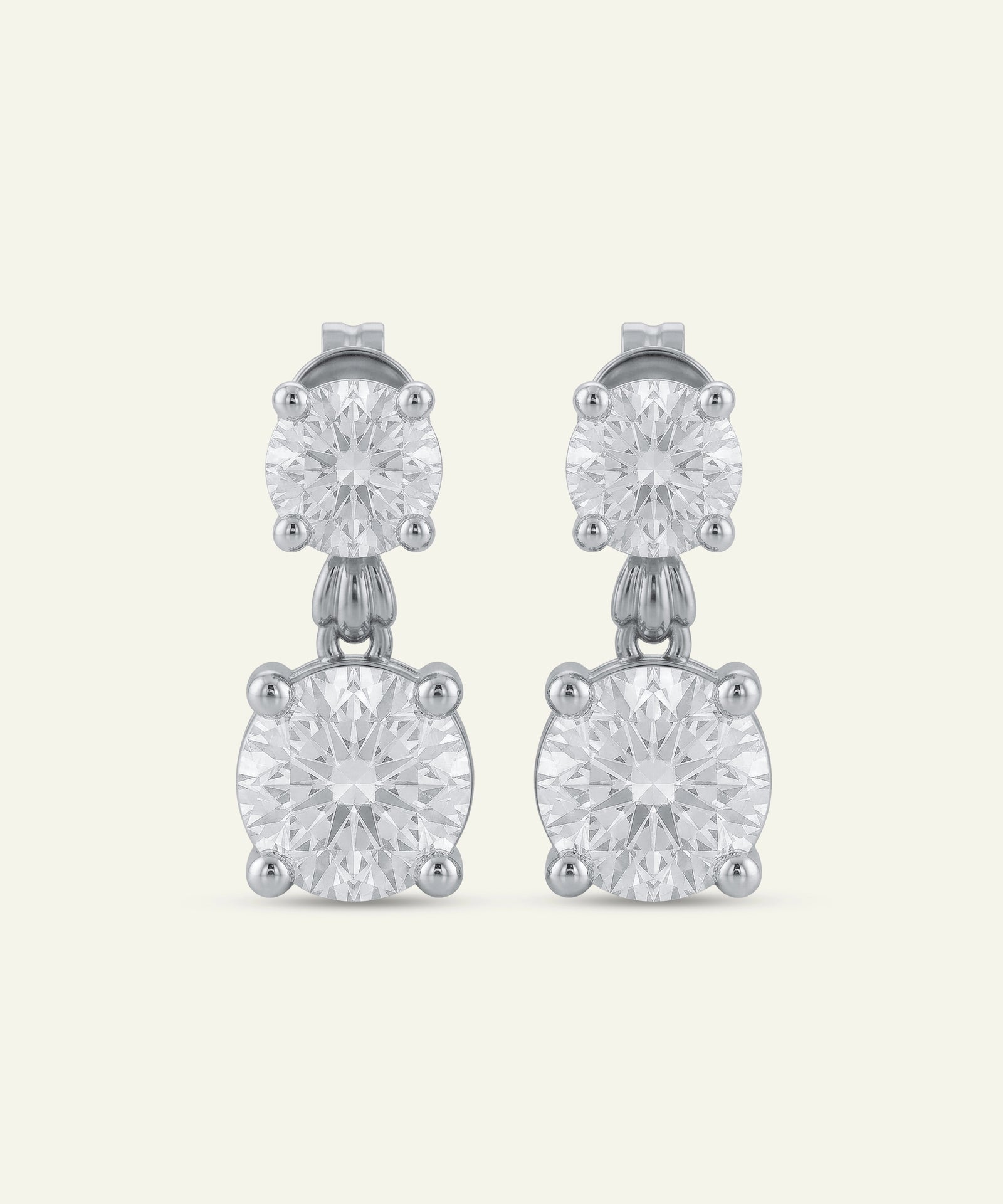 Pair of round brilliant-cut diamond drop earrings in white metal, featuring a smaller round diamond stud connected to a larger round diamond drop in four-prong settings. The earrings are displayed against a light neutral background.