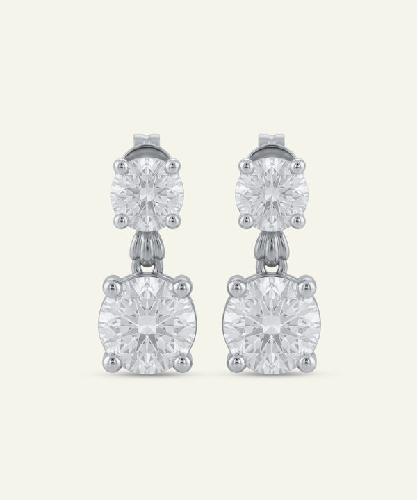Pair of round brilliant-cut diamond drop earrings in white metal, featuring a smaller round diamond stud connected to a larger round diamond drop in four-prong settings. The earrings are displayed against a light neutral background.