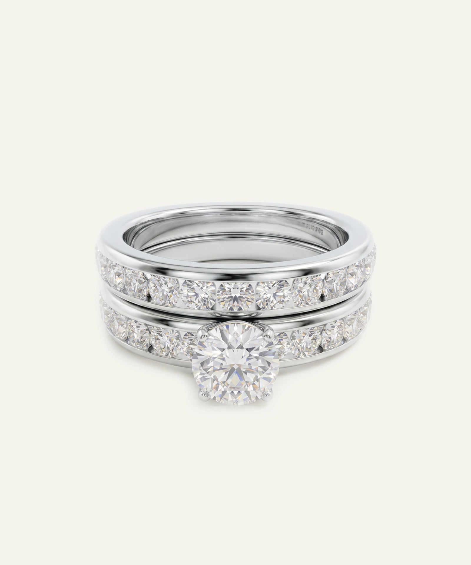 A three-band diamond ring set in polished silver-toned metal, featuring a round-cut center stone on the middle band, with pavé diamond bands above and below, shown on a light cream background.