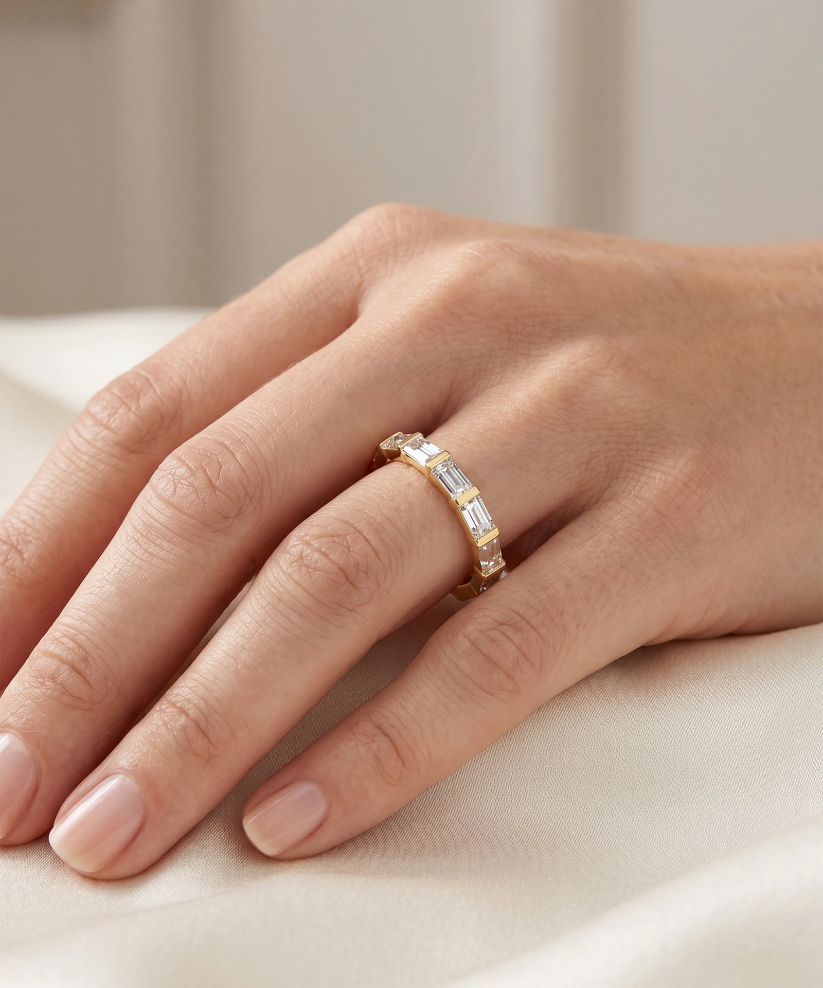 A close-up of a hand wearing a delicate gold band ring set with baguette-cut clear stones, resting on a soft cream-colored fabric.