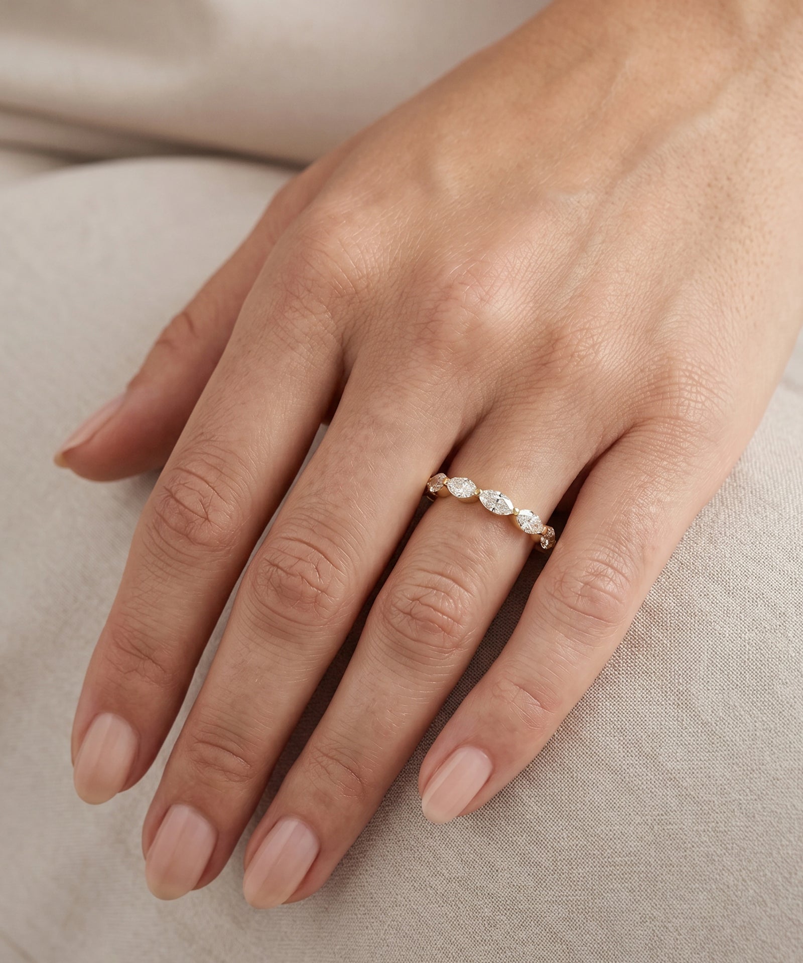 A close-up of a hand wearing a delicate silver band ring with three small clear marquise-cut gemstones, resting on a soft beige fabric surface.
