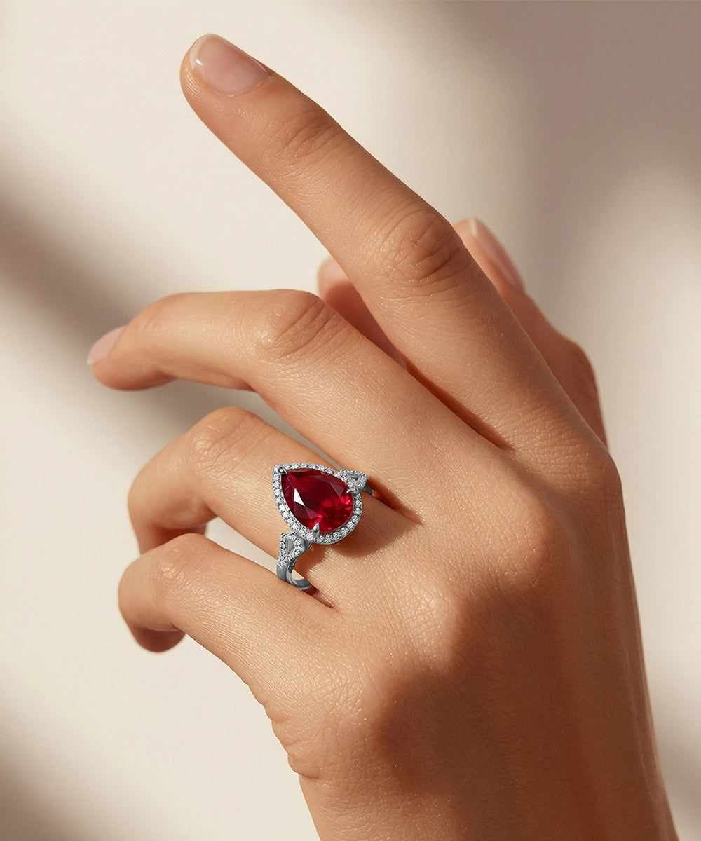 Hand wearing a ring with a pear-shaped red gemstone on a neutral background
