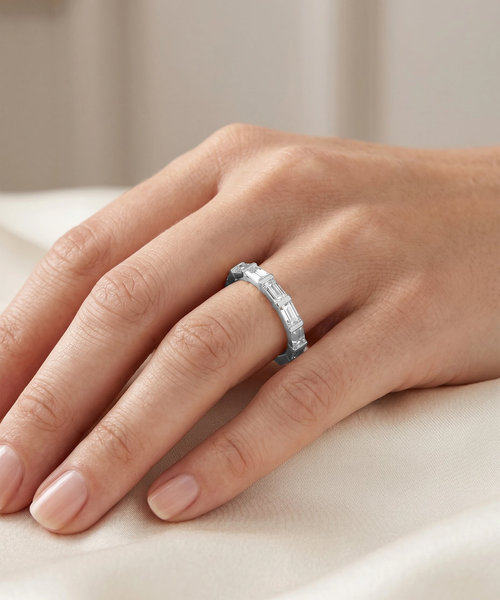 A close-up of a hand wearing a silver band ring with a row of small clear baguette-cut stones, resting on a soft cream fabric surface.