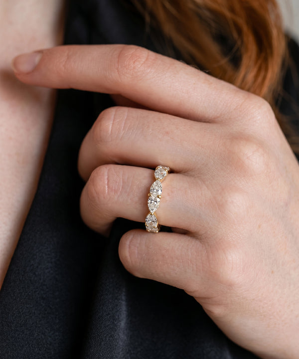 A close-up of a person's hand wearing a delicate gold ring with a row of clear marquise-cut gemstones, set against a dark black fabric background.