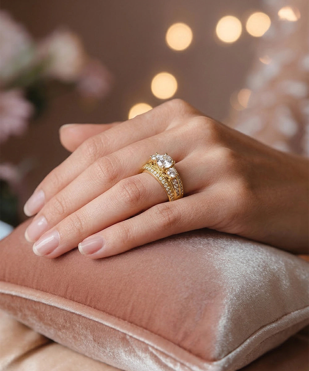Hand wearing a gold ring with a diamond on a pink cushion with blurred lights in the background