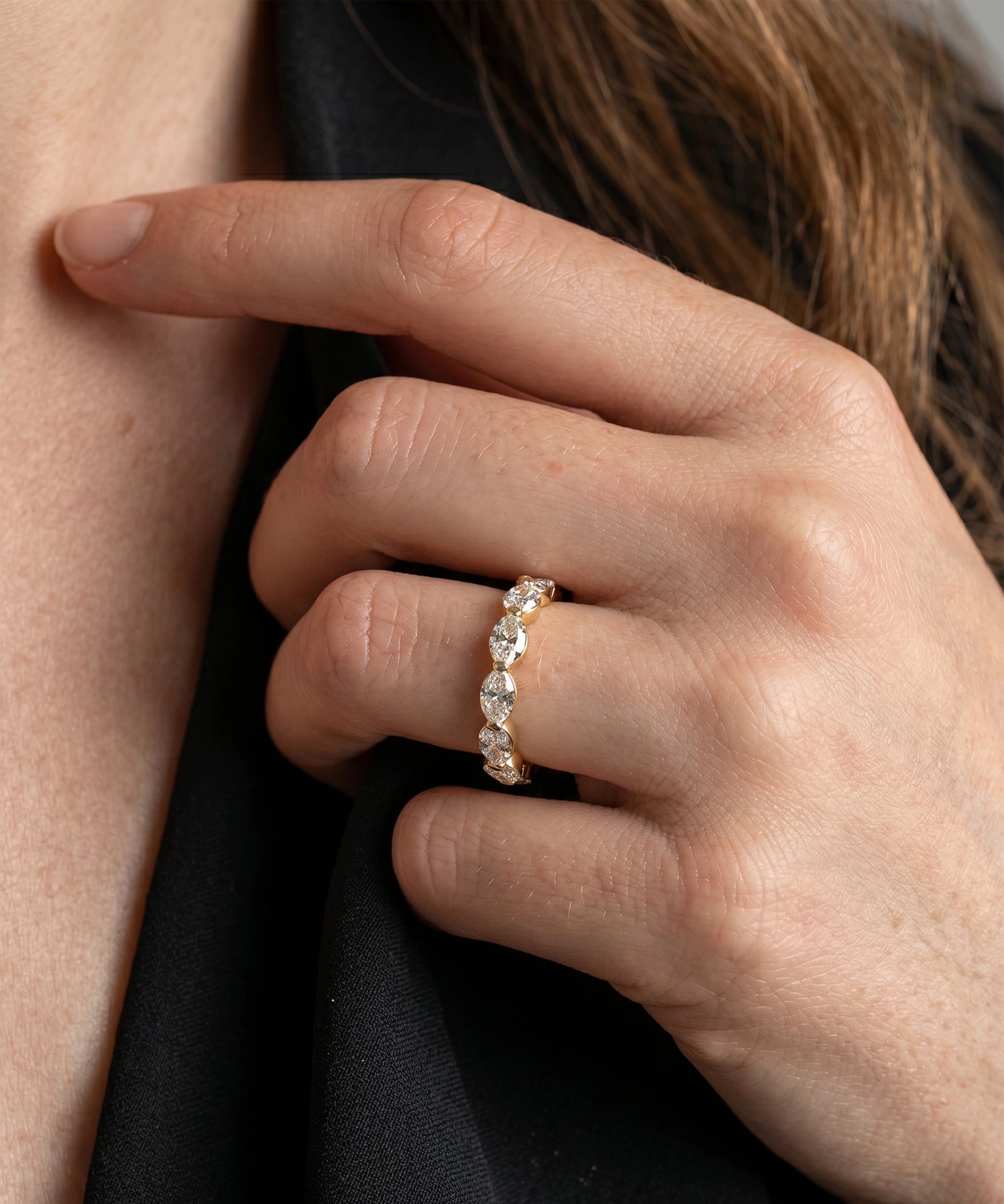 A close-up of a person's hand wearing a delicate gold band ring set with small clear stones, against dark clothing and skin.