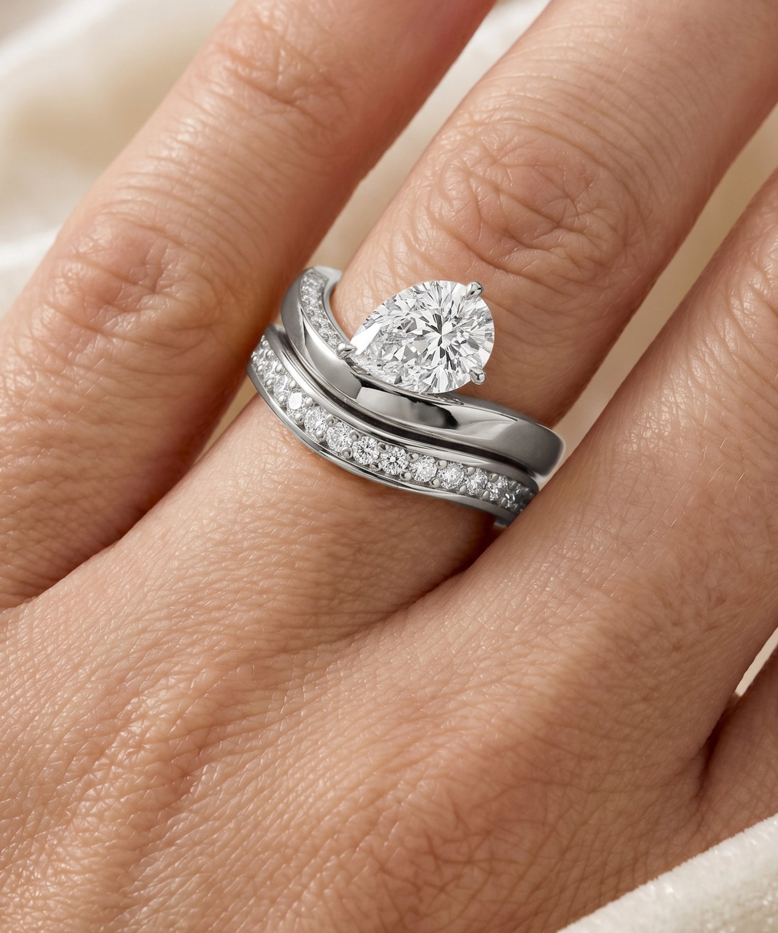 A close-up of a hand wearing a silver-toned wedding band set featuring a pear-cut diamond solitaire ring and a matching pavé band.