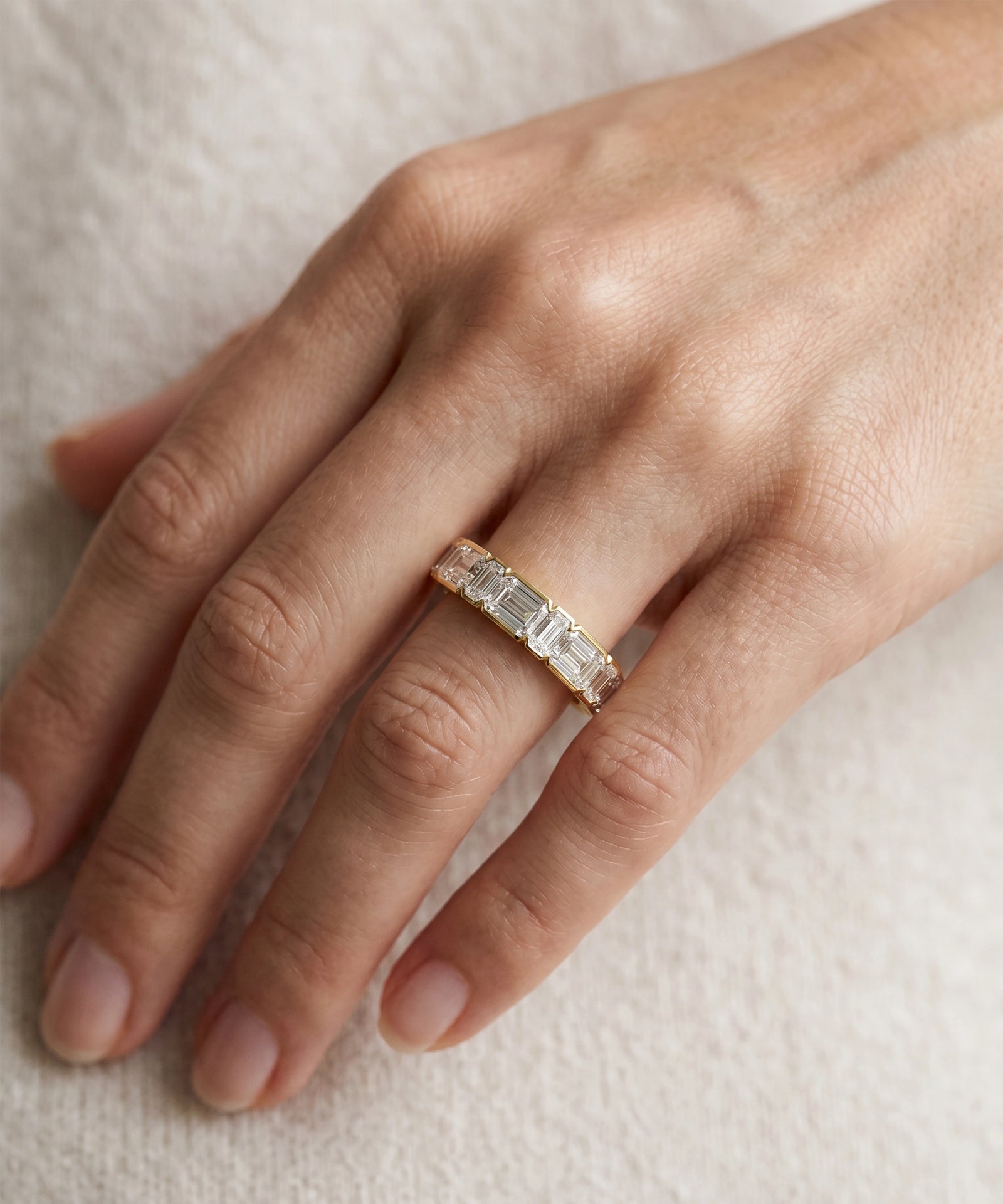 A close-up of a hand wearing a gold eternity ring set with baguette-cut clear stones, resting on a soft, textured cream fabric background.