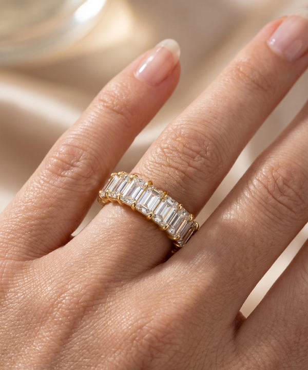 A close-up of a hand wearing a gold eternity ring set with baguette-cut clear stones, set against a soft beige satin background.