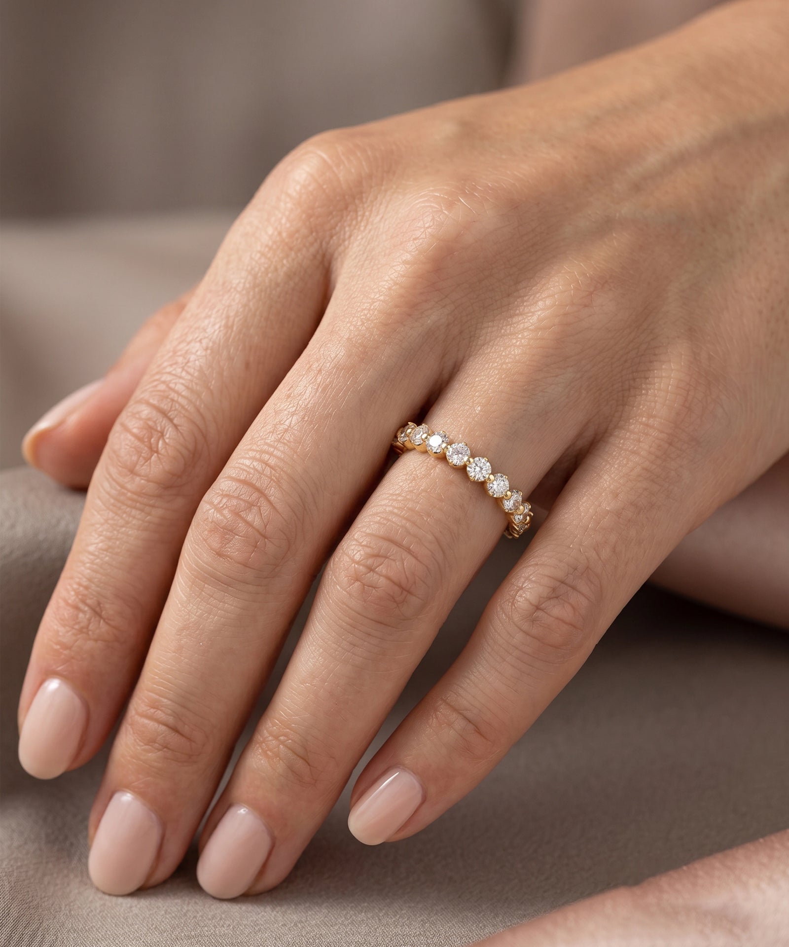 A close-up of a hand wearing a delicate gold band ring set with small clear stones, resting on a soft neutral fabric background.