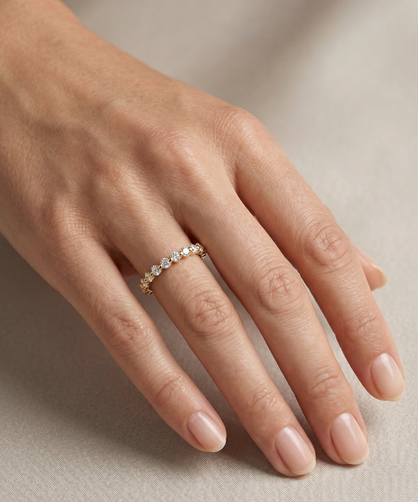 A close-up of a hand wearing a delicate gold ring set with small clear stones, photographed against a soft neutral beige background.