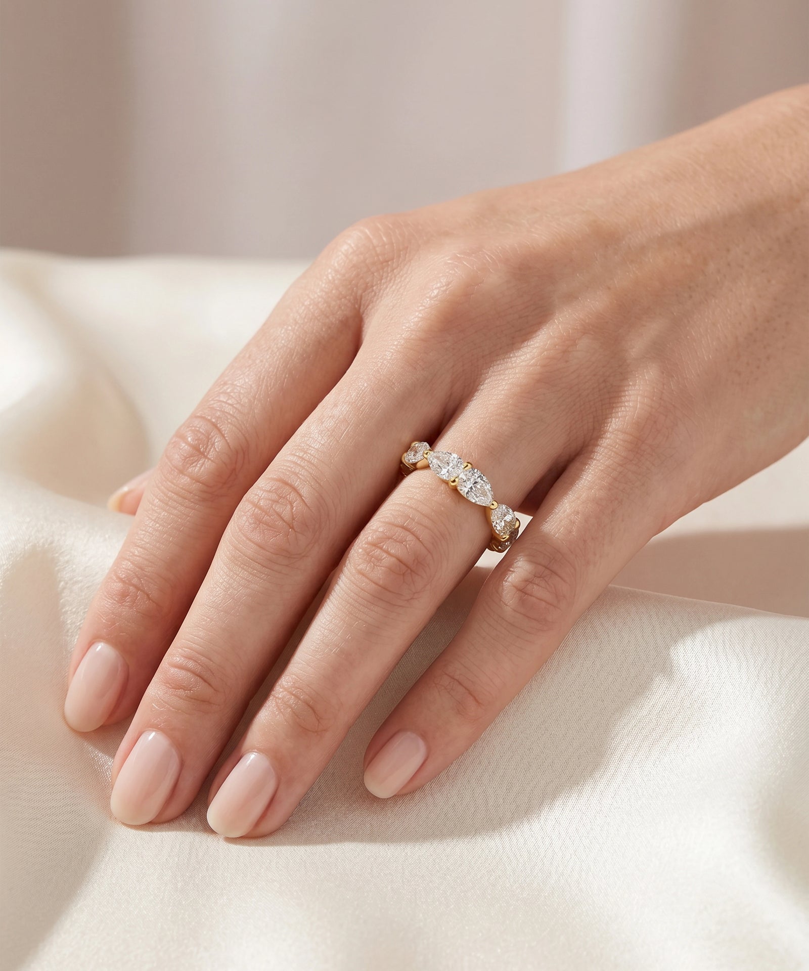 A close-up of a hand wearing a delicate gold ring with a row of small clear stones, resting on soft white fabric.