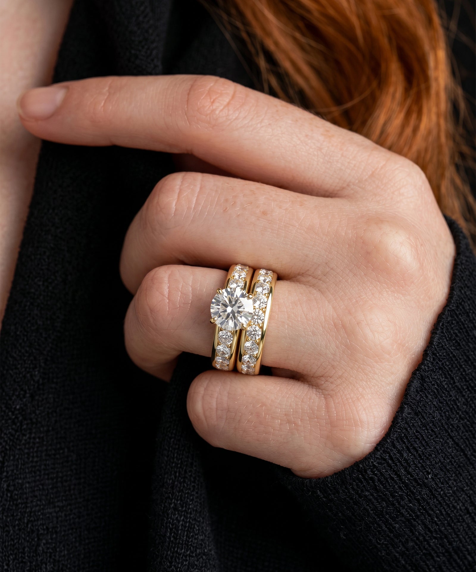 A close-up of a person's hand wearing a gold wedding band with a large round diamond and a matching diamond-encrusted band.