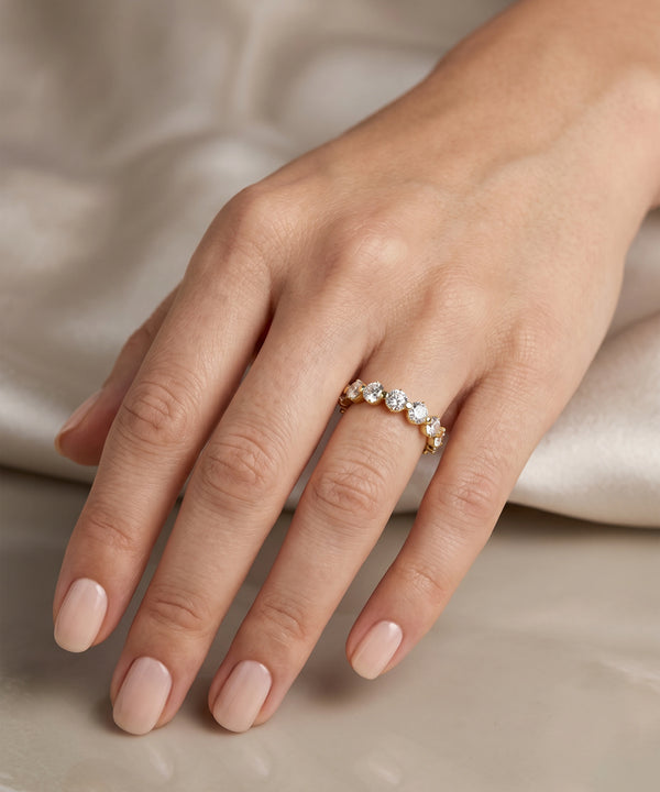 A close-up of a hand wearing a delicate gold ring adorned with small clear gemstones, set against a soft beige satin background.