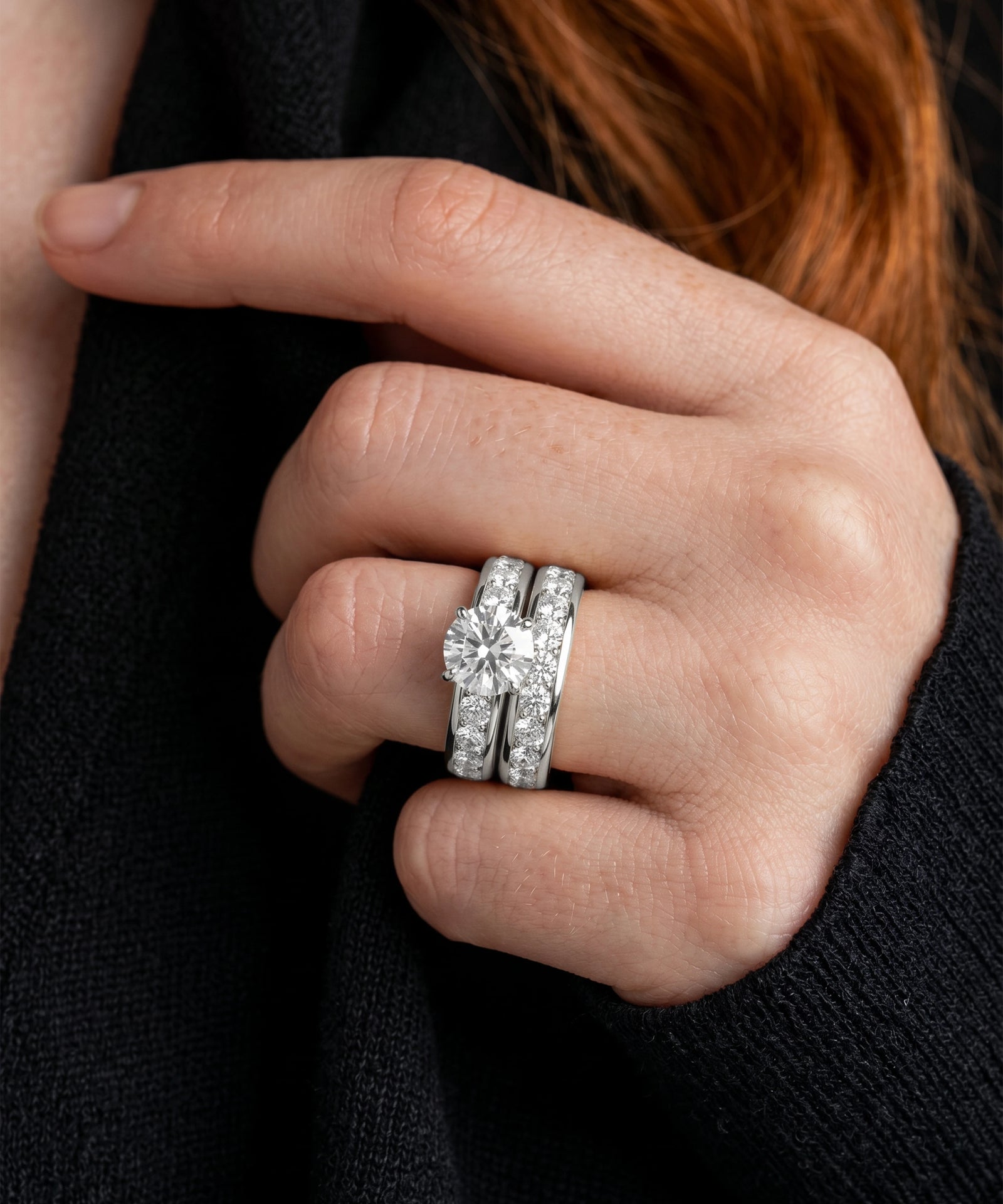 A close-up of a hand wearing a silver-toned wedding band set with a large round-cut center stone and smaller pavé-set stones, worn over a black knit top.