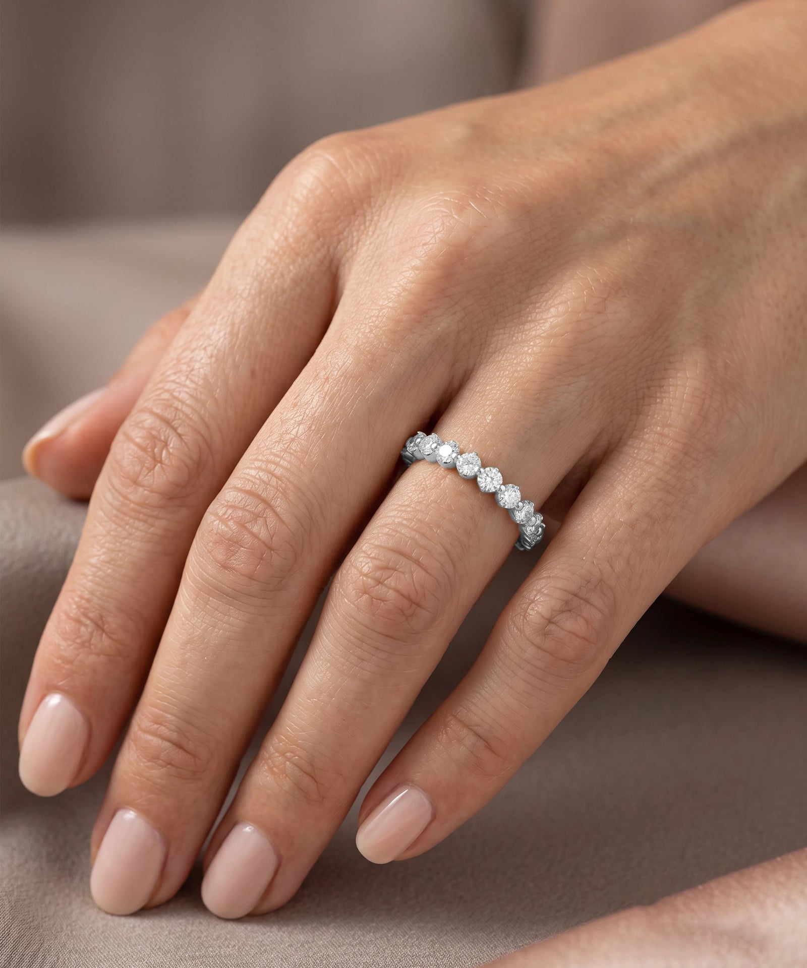 A close-up of a hand wearing a delicate silver band ring set with small round clear stones, resting on a soft beige fabric background.