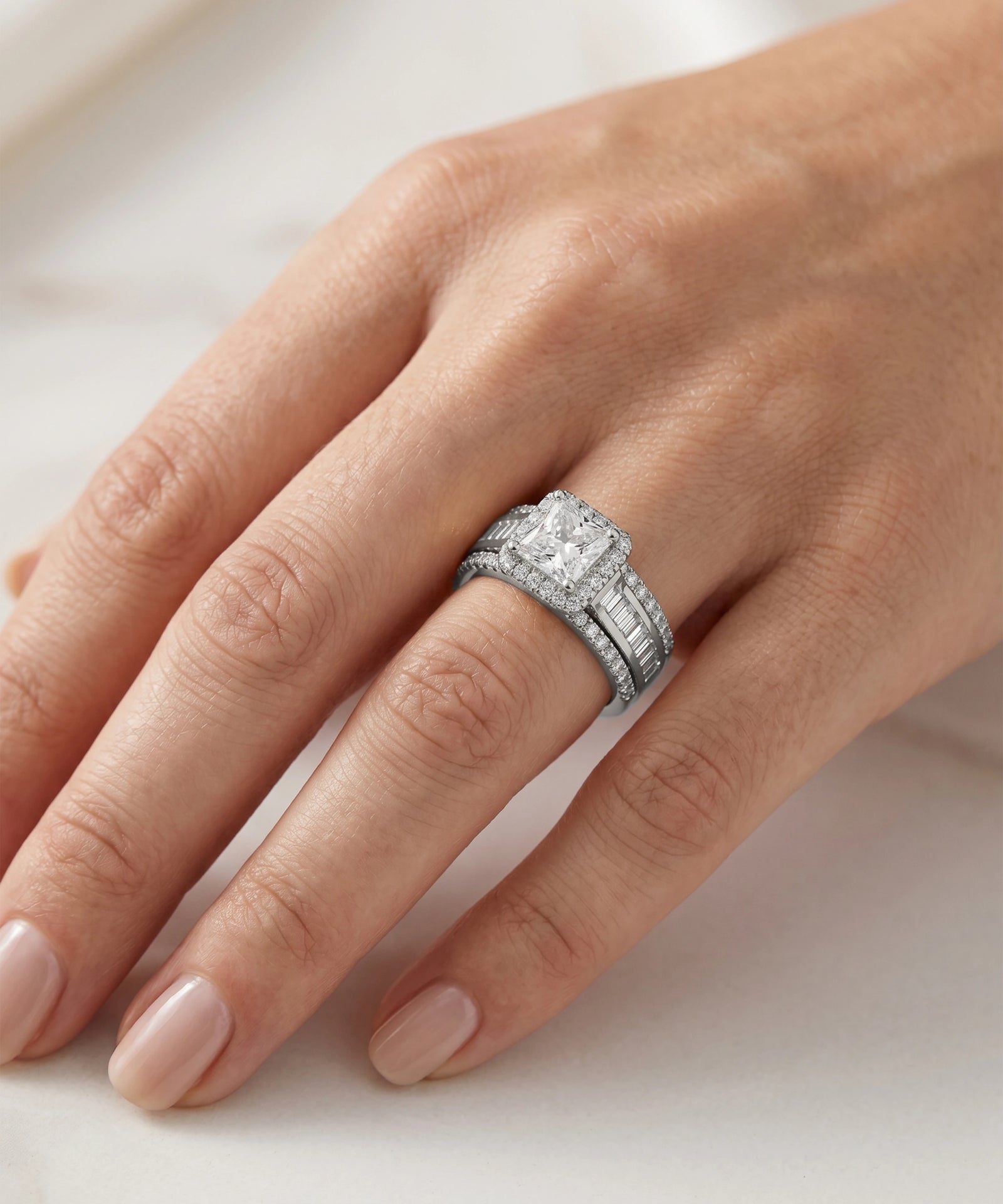 A close-up of a hand wearing a silver-toned wedding band with a large rectangular diamond center stone and smaller diamonds along the band, set against a soft light background.