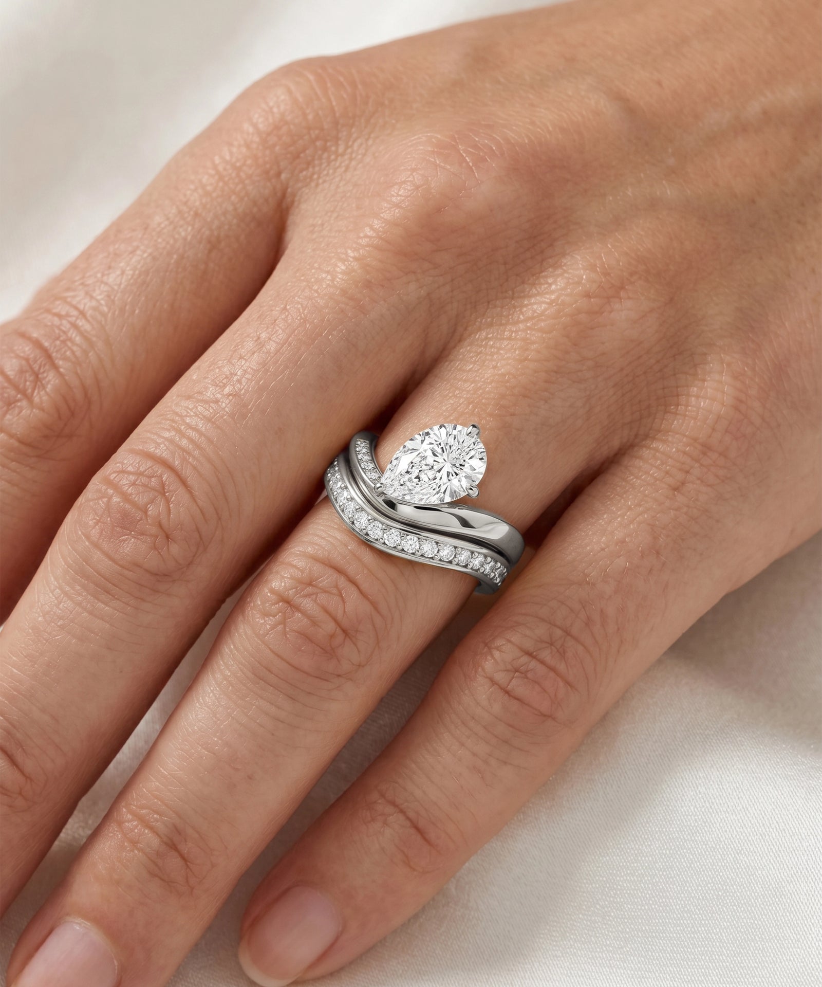 A close-up of a hand wearing a silver-toned wedding band set with a pear-cut diamond center stone and a smaller diamond-accented band.